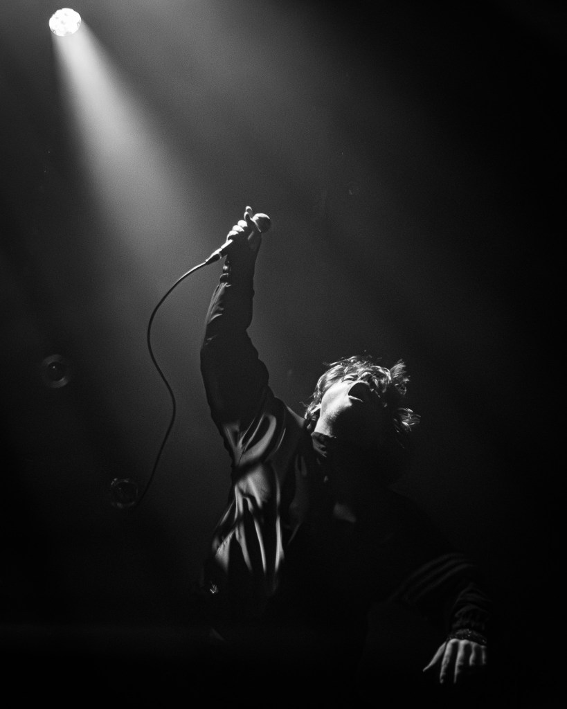 Black and white photograph of a singer performing on stage, holding a microphone and reaching towards the spotlight