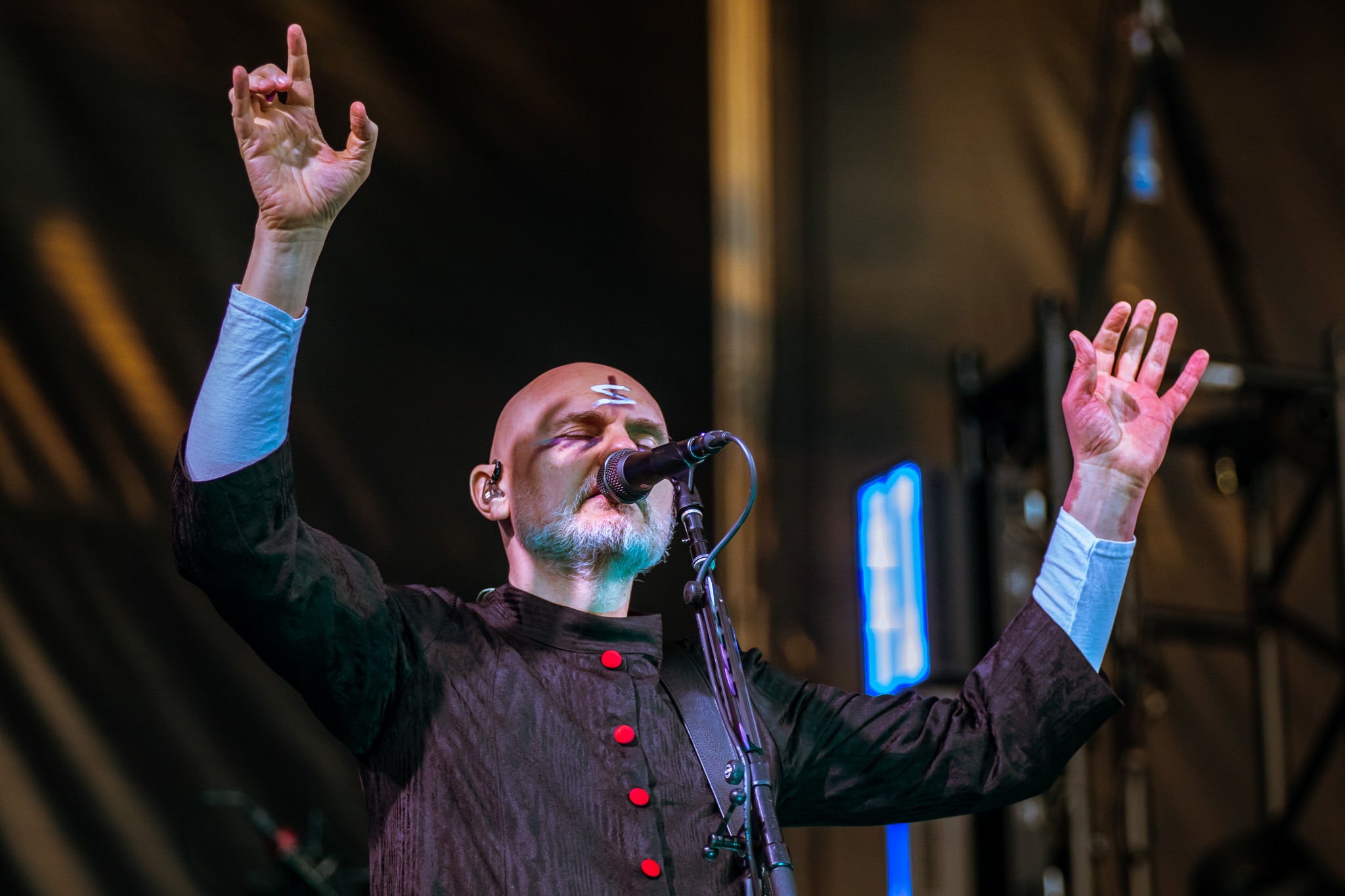 Billy Corgan on stage with raised hands, passionately performing into a microphone. The musician has a shaved head and wears a dark outfit with bright buttons