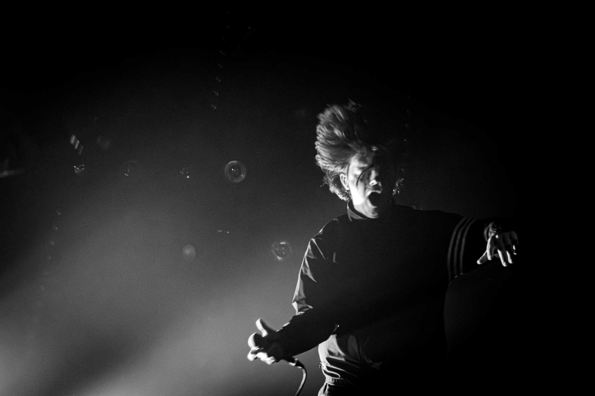A black and white image of a vocalist on stage. The artist's hair is tousled, and they are captured in a dynamic pose under dramatic lighting