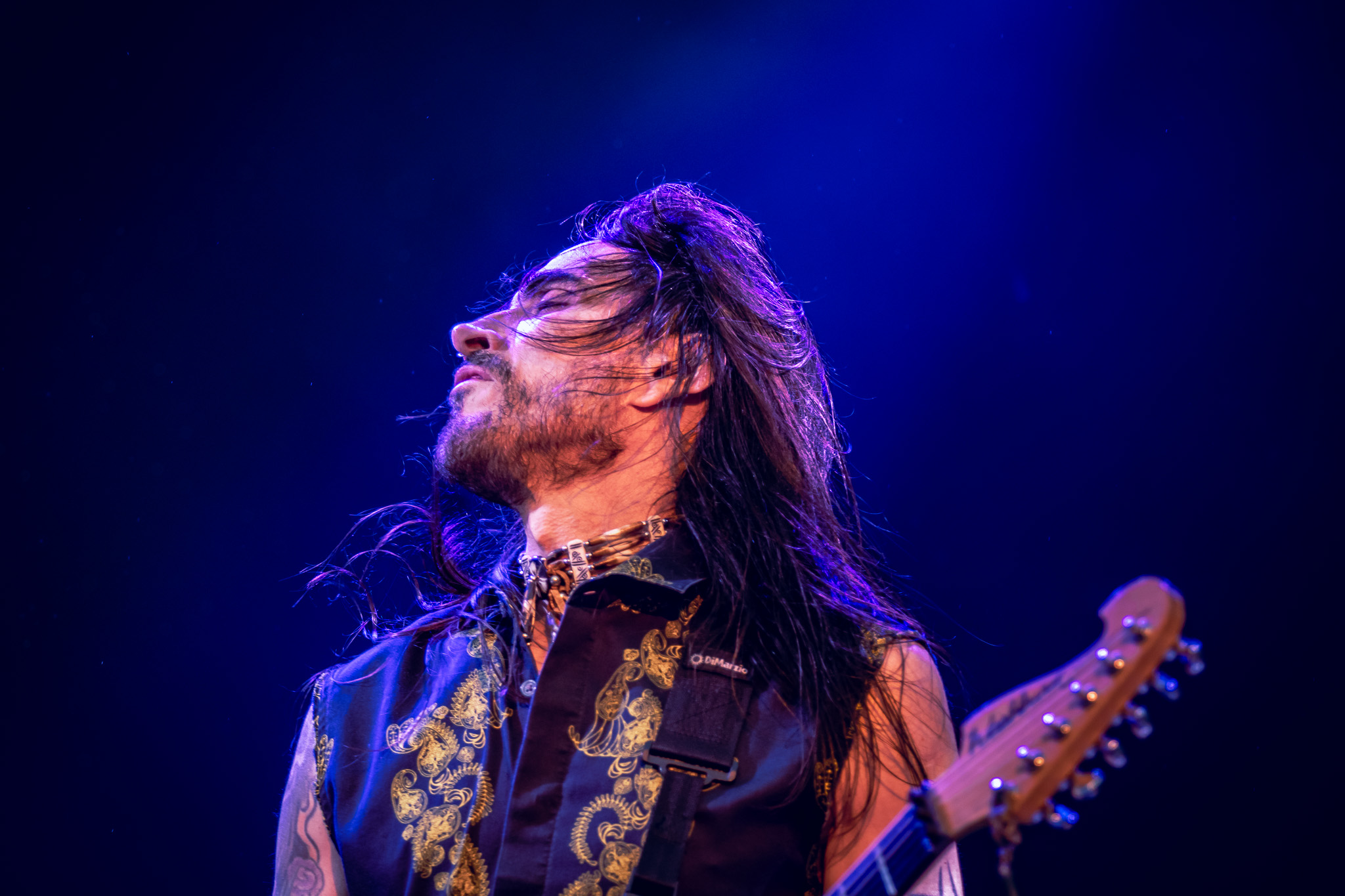 Guitarist Nuno Bettencourt perrforming on stage, illuminated by cool blue lights. The musician is captured mid-performance, with flowing long hair and a confident expression