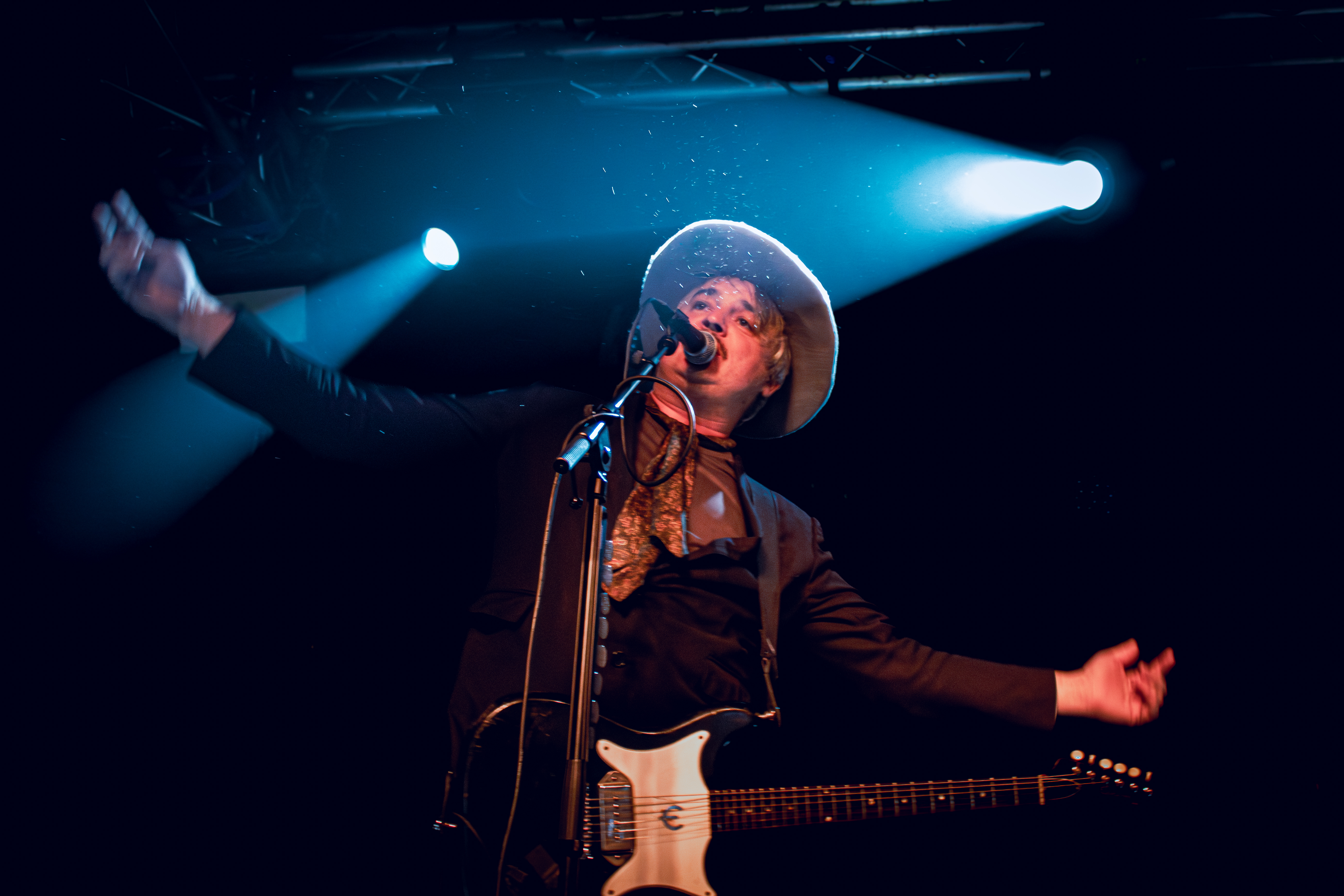 Pete Doherty wearing a hat passionately performing on stage with an electric guitar, illuminated by stage lights.