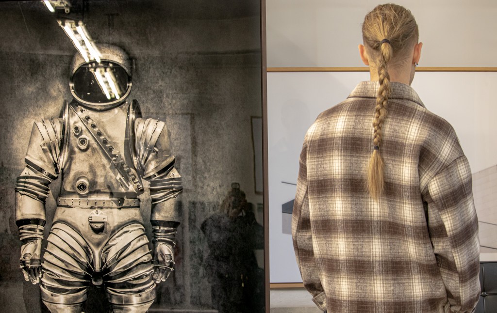 n astronaut suit displayed in an exhibit, with a visitor in a plaid shirt and braided hair observing from the side