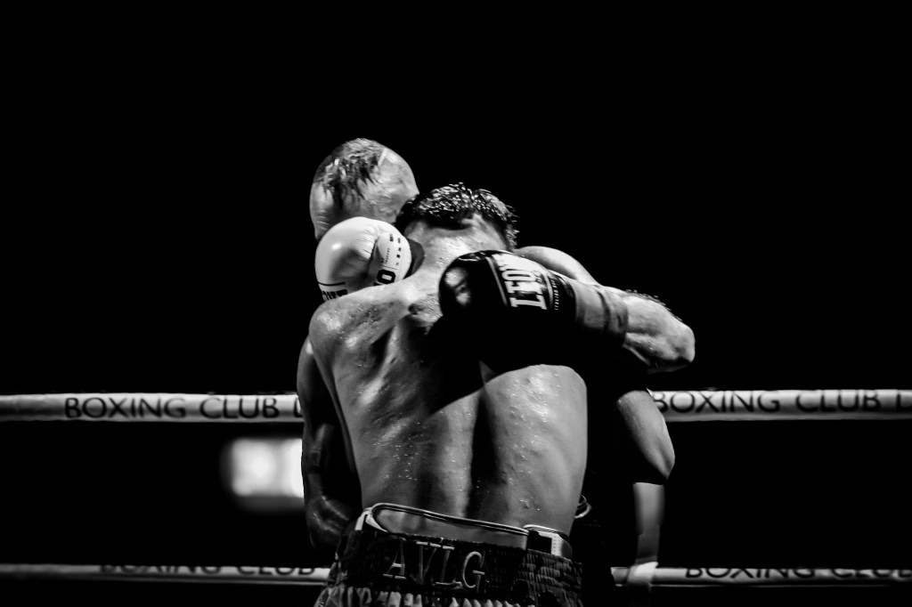 A black and white image of two boxers in a clinch during a match. One boxer has his back to the camera while the other leans in.