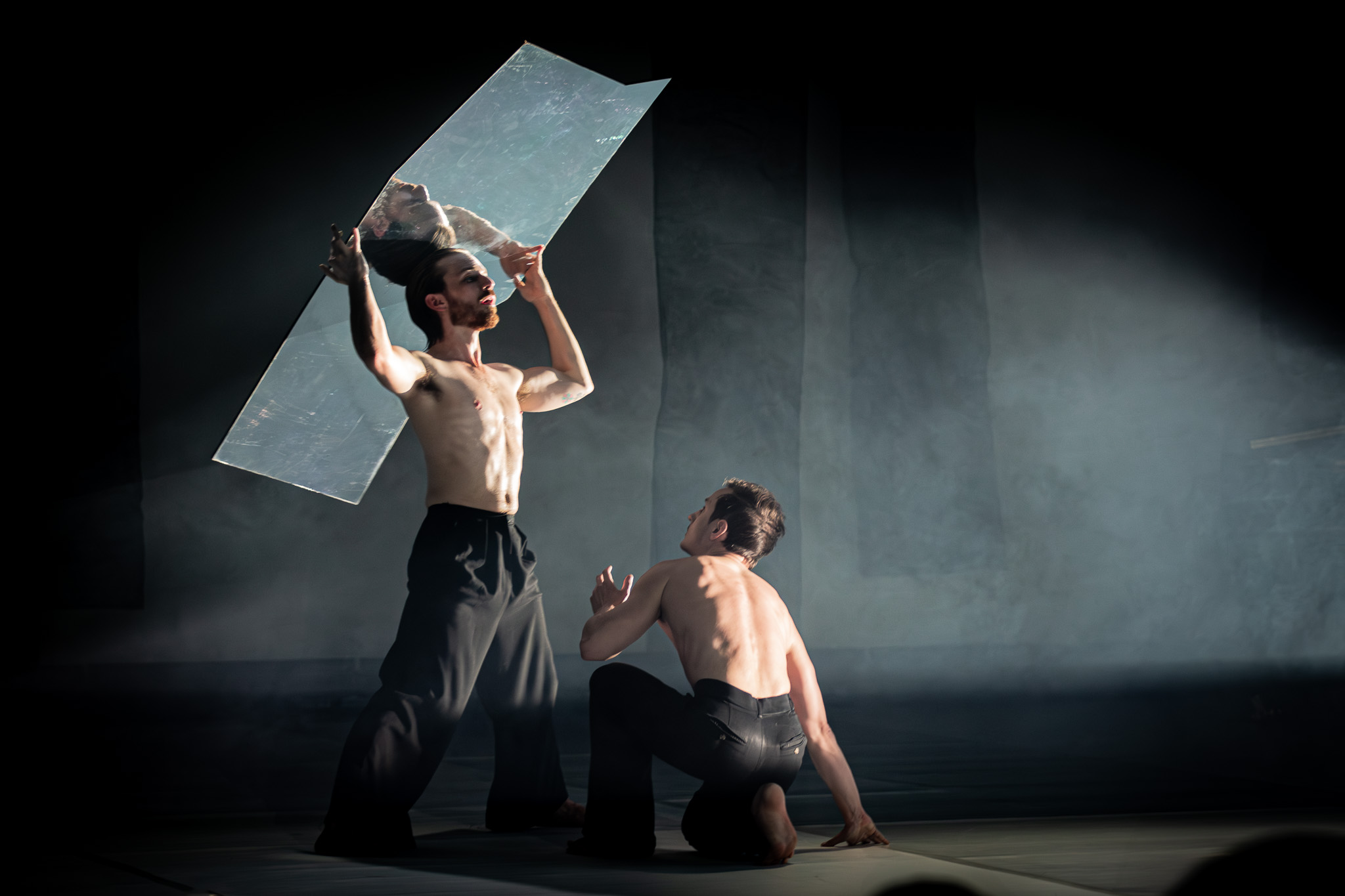 Two male dancers: one dancer holds a mirror above his head, reflecting light while the other is kneeling in a dramatic pose.