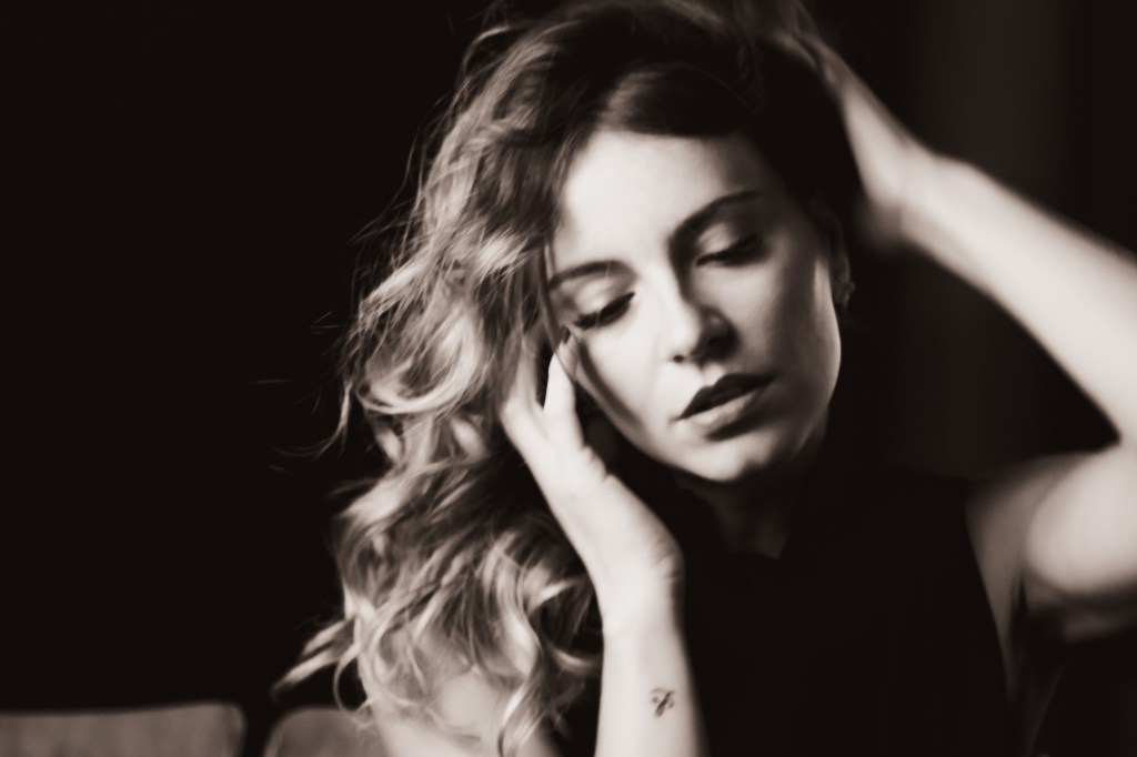 A black and white portrait of a woman with wavy hair, gently holding her head with both hands. Her eyes are closed, conveying a sense of introspection and emotion.