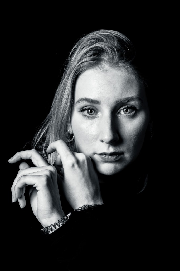 Black and white portrait of a woman with long hair, gazing directly at the camera with hands delicately positioned near her face against a dark background.