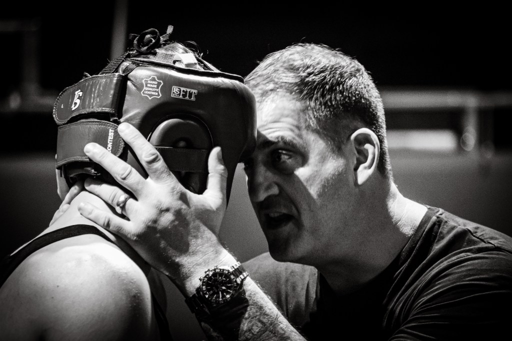 Black and white image of a boxing coach intensely speaking to a boxer wearing protective headgear, capturing a moment of guidance and motivation before a match.