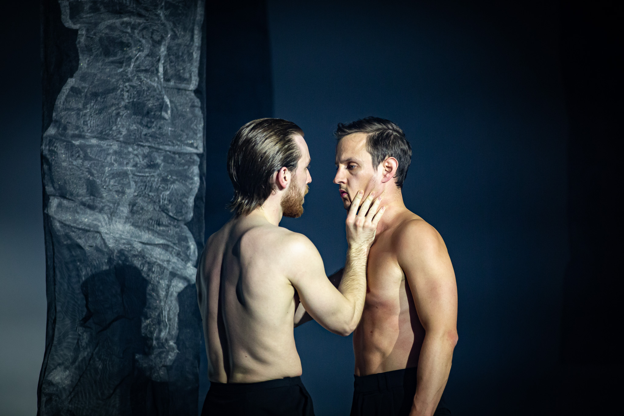Two male dancers, both shirtless, share an intimate moment on stage. One dancer gently touches the other's face.