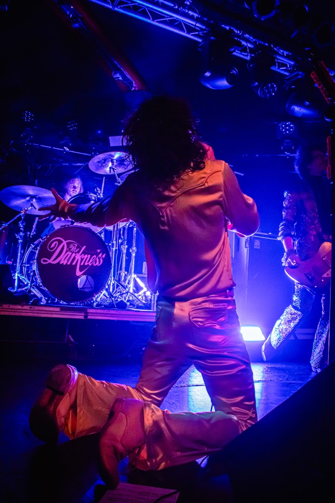 Justin Hawkins kneeling on stage with his back toward the audience