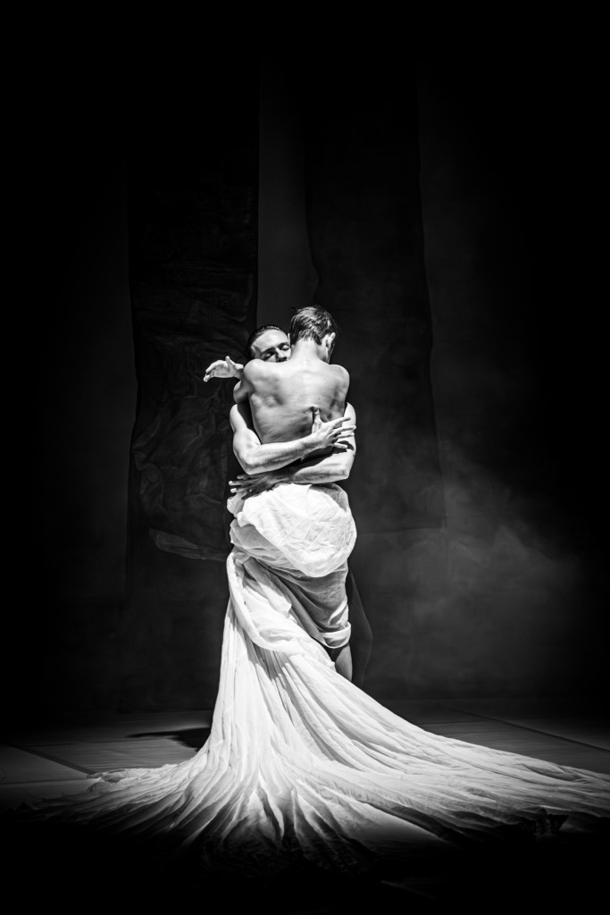  Two dancers embrace tightly, wrapped in flowing fabric that trails behind them.