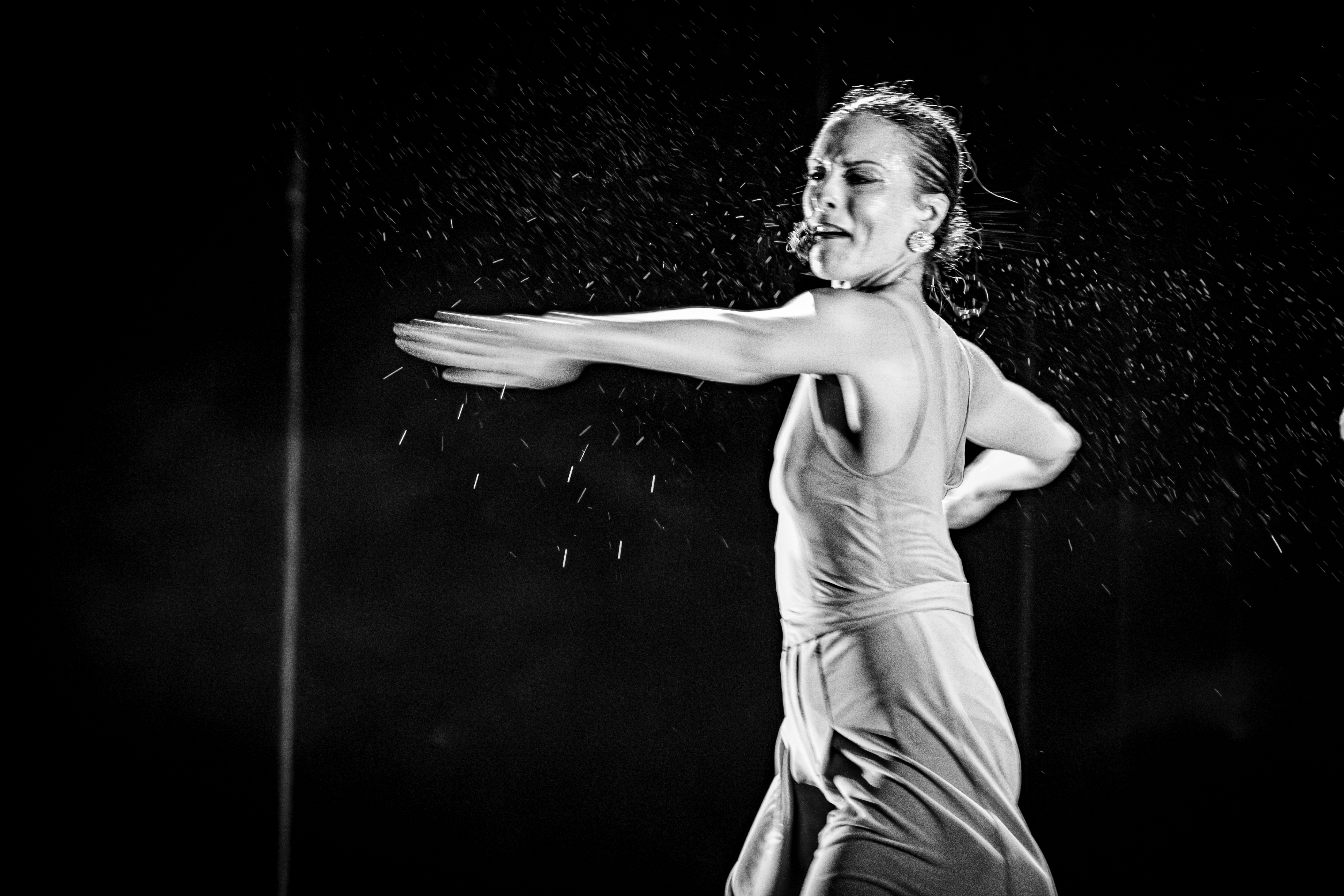 A female dancer performing on stage, captured in black and white. Water droplets are visible, creating a sense of movement as she extends her arms.