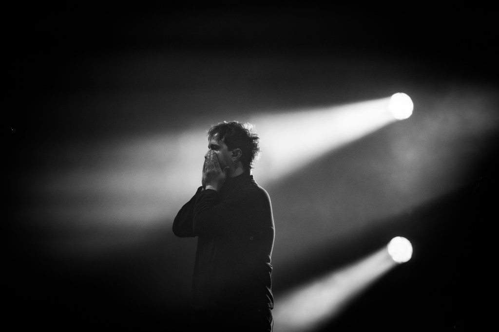 lead singer standing in profile with his hands covering his face, illuminated by soft light beams in a black and white setting.