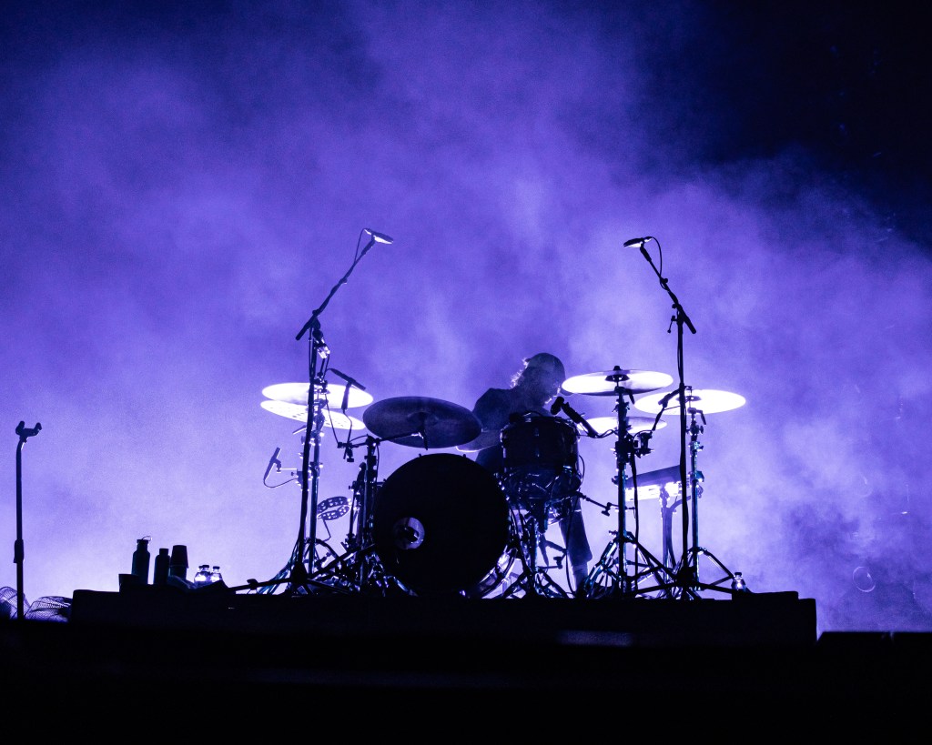 silhouette of a drummer performing on stage, surrounded by a misty purple atmosphere, with drumsticks raised and illuminated by subtle lighting.