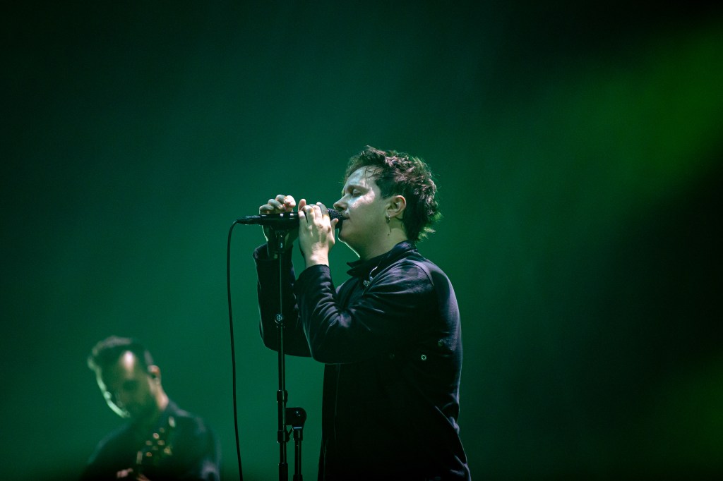 lead singer passionately performing into a microphone, bathed in green lighting, with a guitarist partially visible in the background.