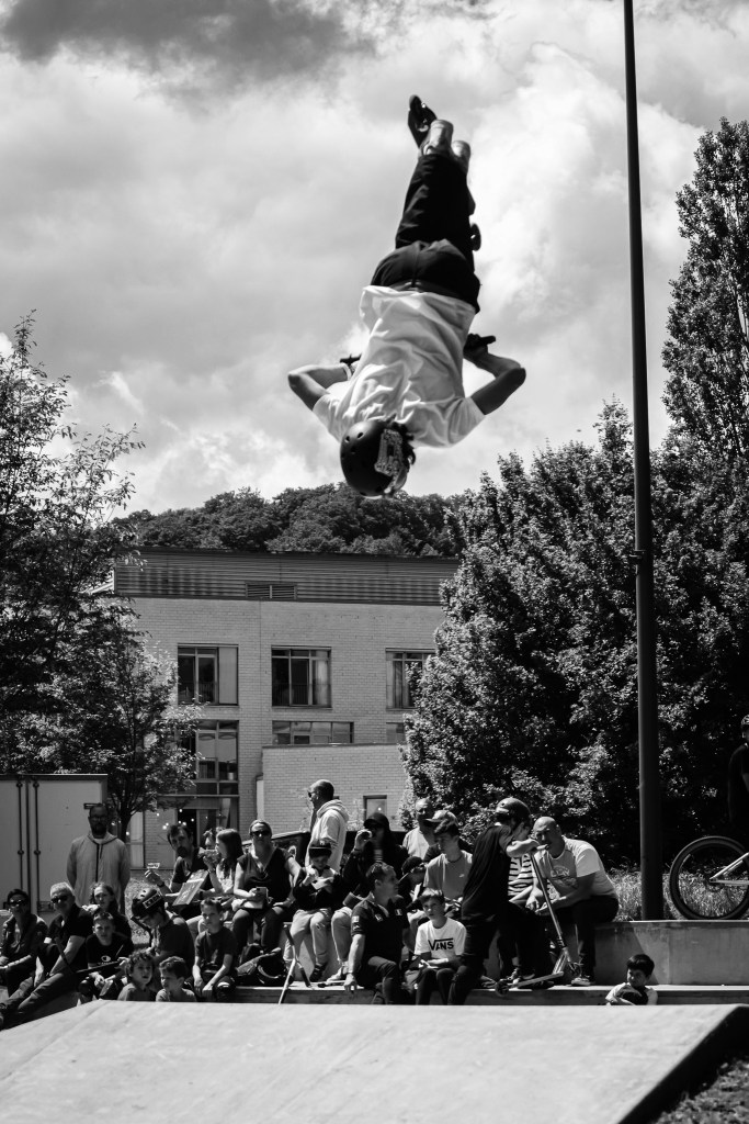 Black and white image of a scooter rider performing a spectacular flip mid-air, with an audience watching in awe at a skatepark,