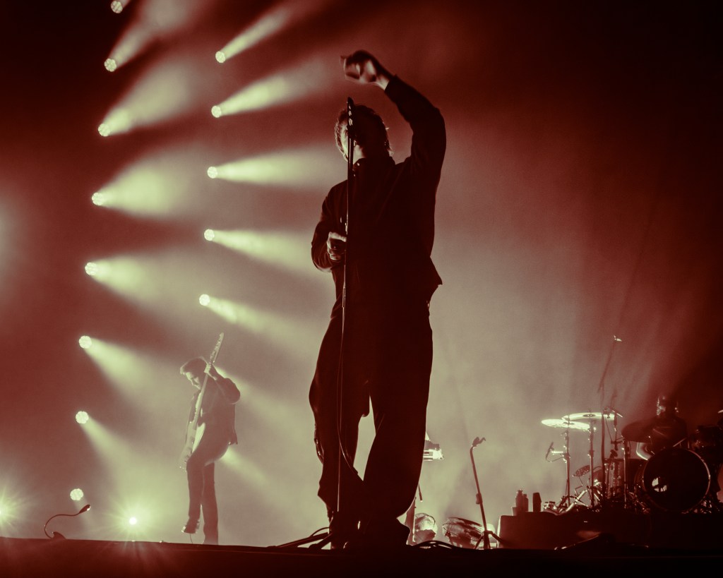 A silhouette of a lead singer with an outstretched arm on stage, accompanied by a guitarist in the background, all set against warm, glowing lights.