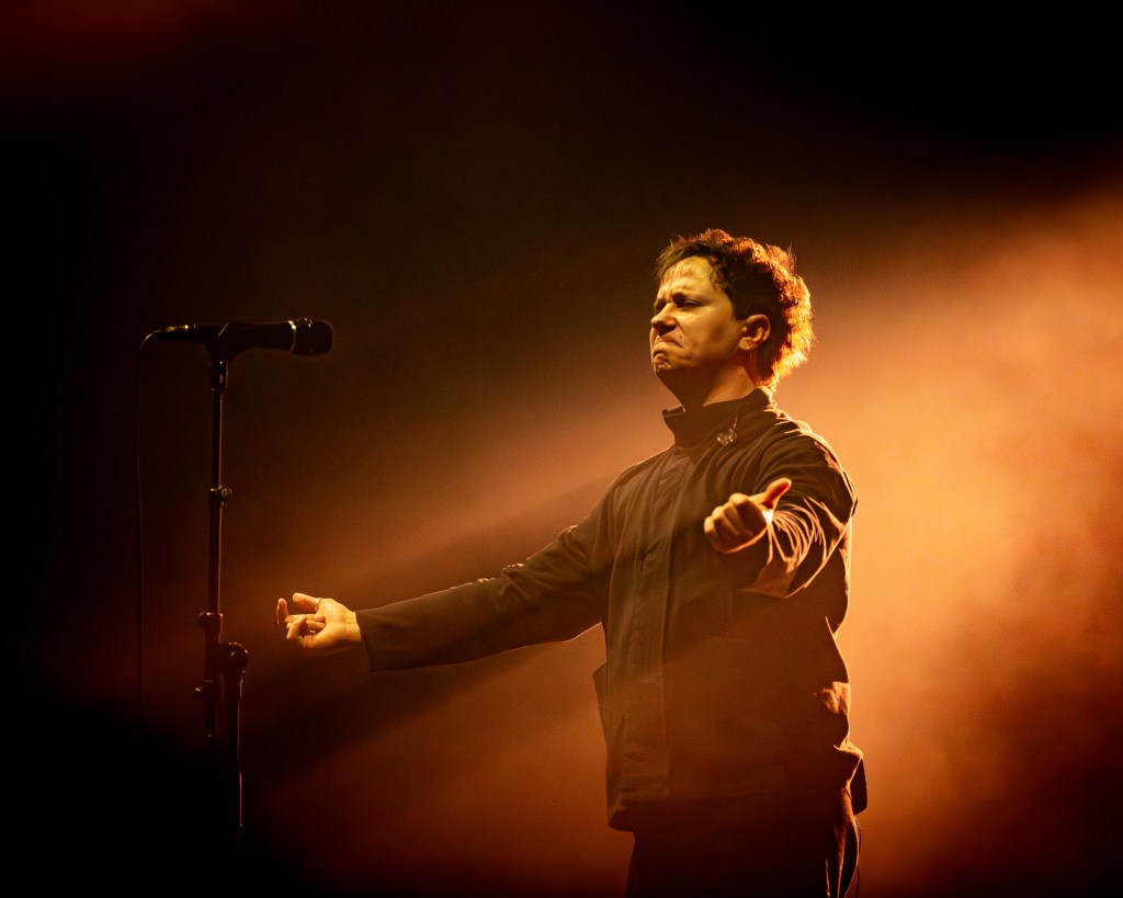 lead singer passionately performing on stage, with an expressive facial expression and arms outstretched, surrounded by warm, dramatic lighting.