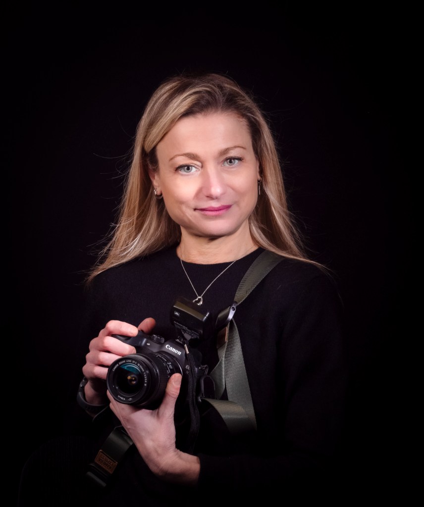 Portrait of a female photographer with long blonde hair, smiling while holding a camera.
