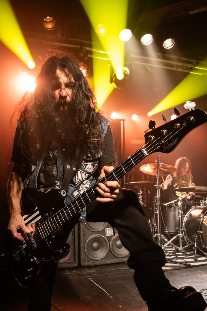 Bassist performing live, intensely focused while playing an electric bass guitar, with vibrant yellow stage lights and a drummer visible in the background