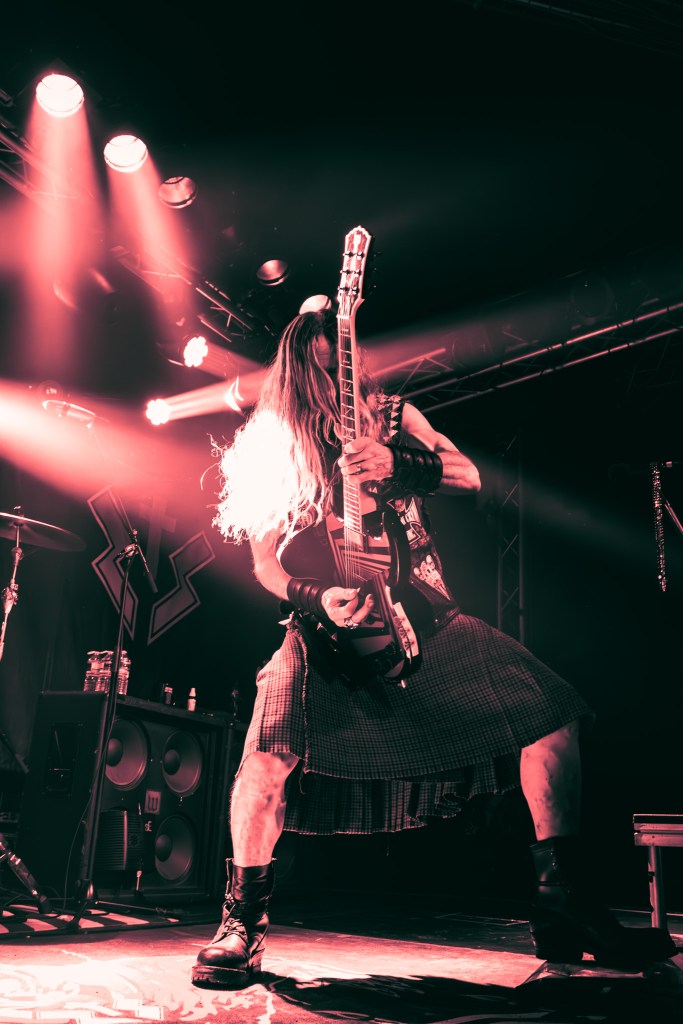 "Zakk Wylde of Zakk Sabbath passionately playing an electric guitar on stage, with dynamic lighting casting vibrant hues,