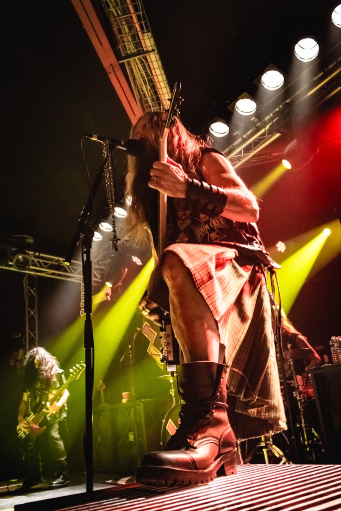Zakk Sabbath guitarist performing on stage under dramatic yellow lighting, wearing a leather vest and kilt