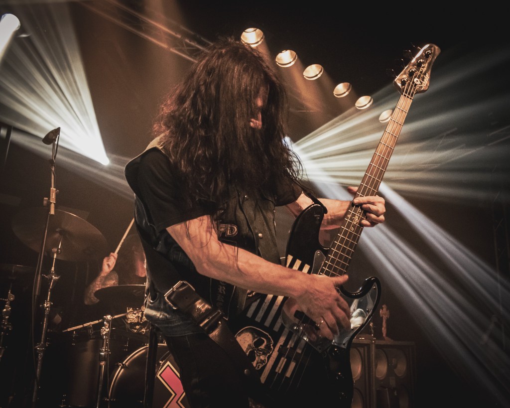 Bassist of Zakk Sabbath performing live, passionately playing an electric bass guitar, with striking light beams illuminating the stage