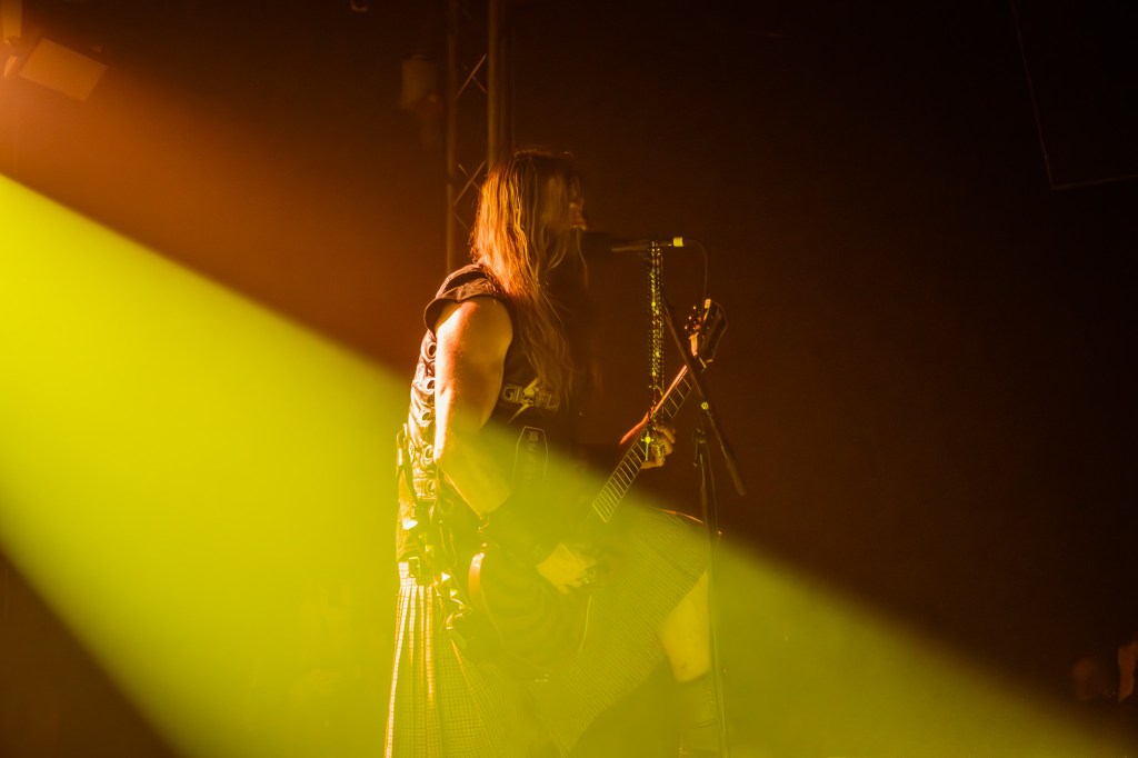 guitarist performing on stage under dramatic yellow lighting, wearing a leather vest and kilt