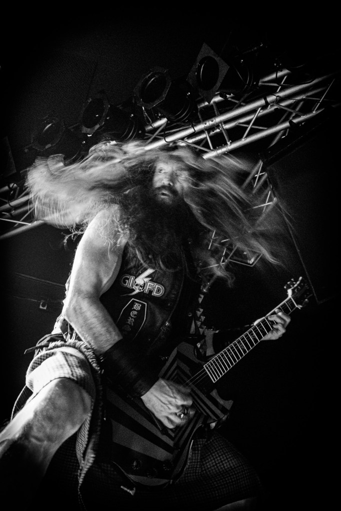 Zakk Wylde performing on stage in a kilt and boots with flowing hair