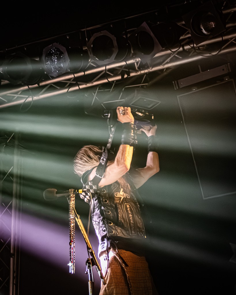 Zakk Wylde raising his electric guitar high above his head during a live performance, surrounded by dramatic beams of stage light