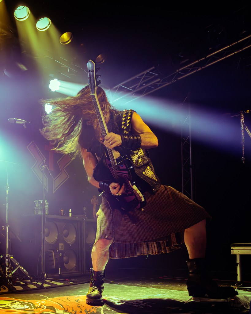 Zakk Wylde performing live showcasing an energetic guitar solo on stage while wearing a kilt and leather vest, under dramatic lighting