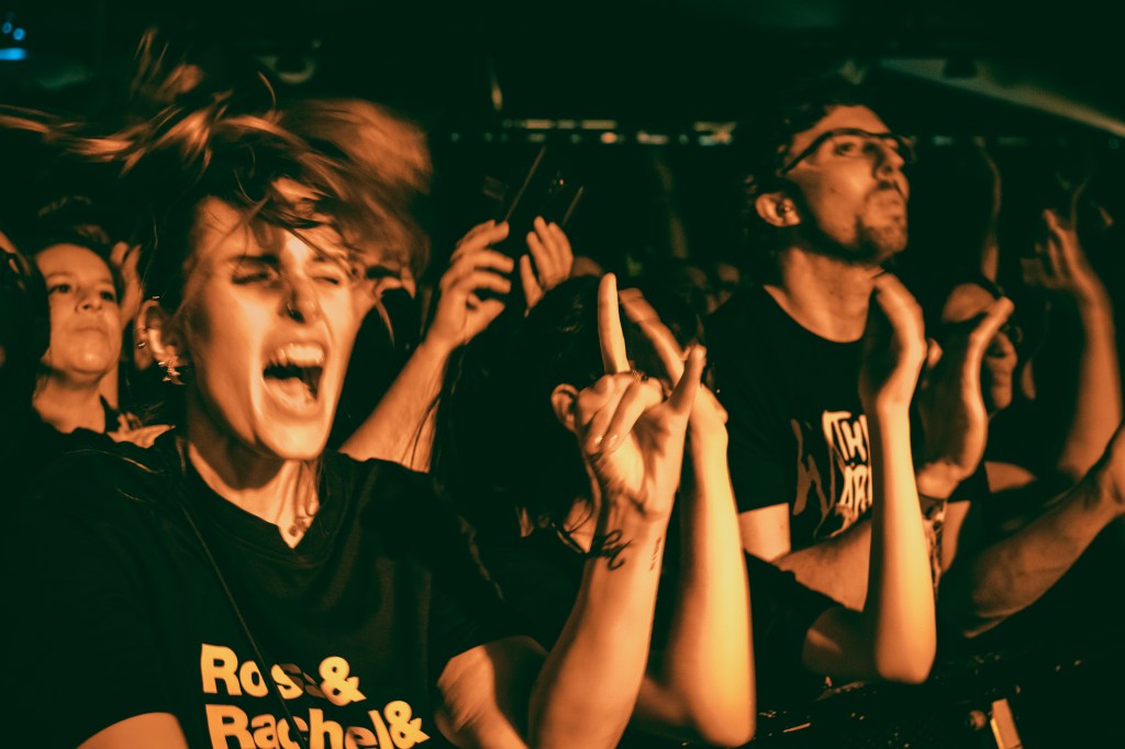 A lively crowd at a concert, with a woman in the foreground passionately expressing excitement and making a rock hand gesture.
