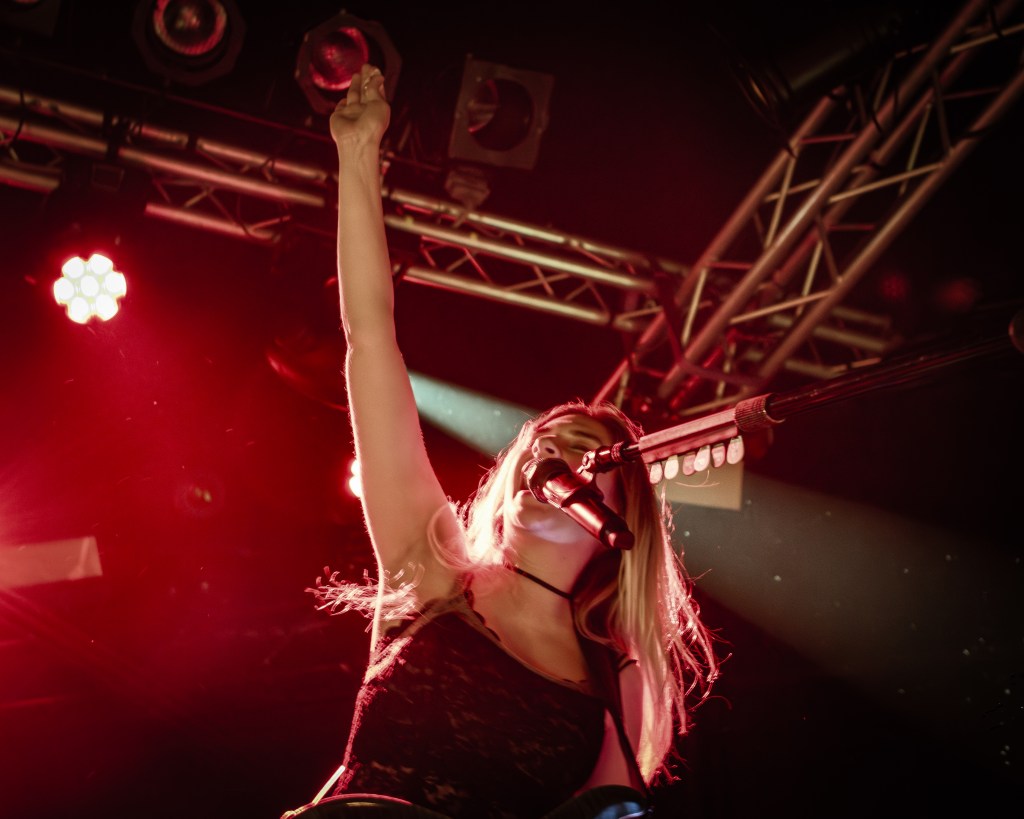A vocalist raising her arm in a powerful gesture during a performance, with vibrant red stage lights creating a dramatic atmosphere. The focus is on her expressive face and dynamic presence on stage.