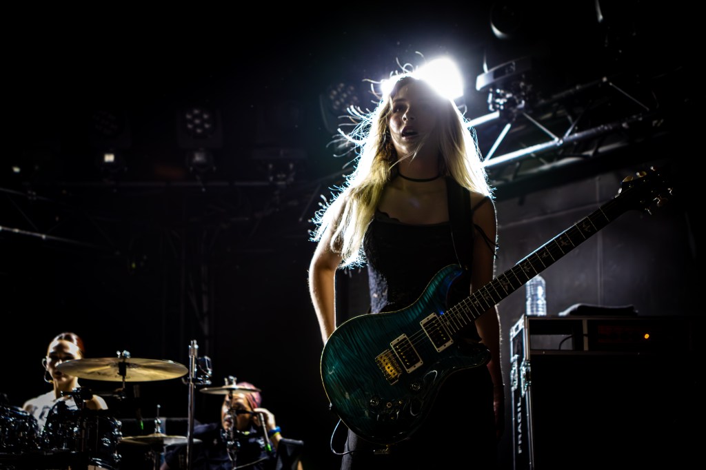 A guitarist on stage, confidently holding her guitar with flowing hair illuminated by bright lights. In the background, a drummer is visible, adding to the dynamic atmosphere of the live performance.