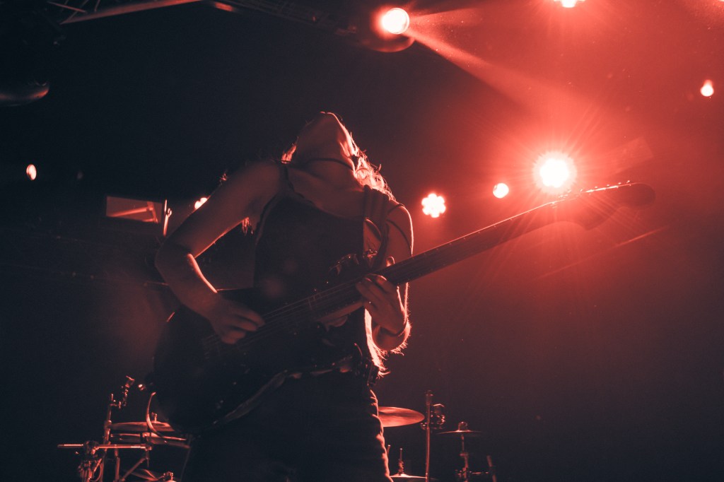 A guitarist in a powerful pose, leaning back as she plays, surrounded by dramatic red stage lights.
