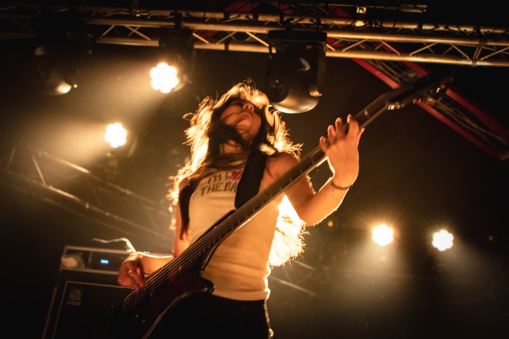 A female bass player performing energetically on stage, with flowing hair and a focused expression. The scene is illuminated by bright stage lights
