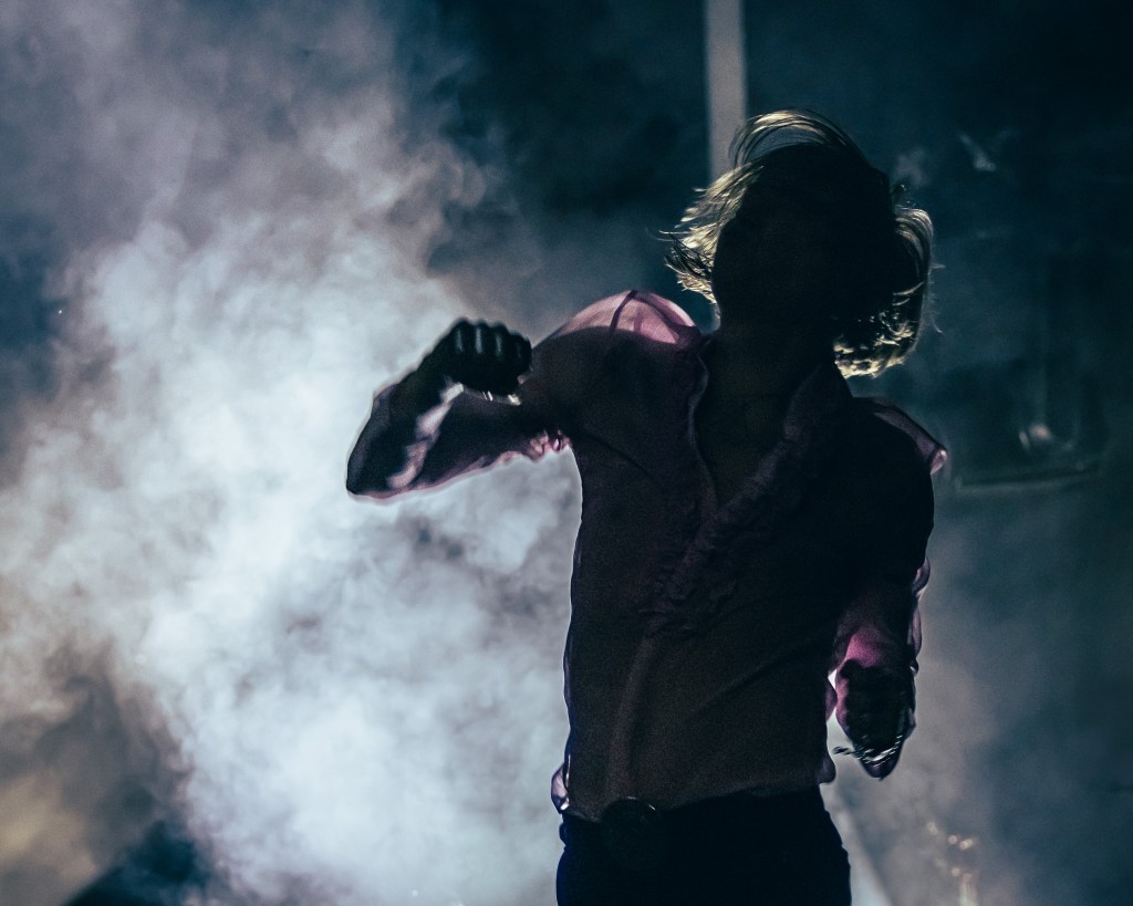 Silhouette of a singer against a smoky backdrop, wearing a ruffled pink shirt, with dynamic hair movement suggesting energy and motion.