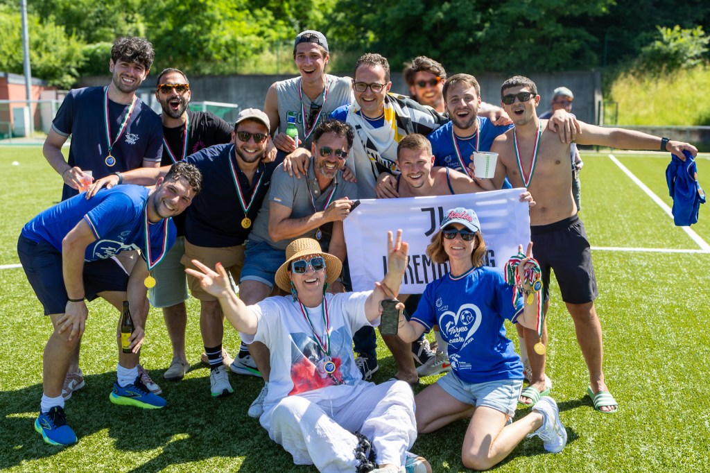 Group of smiling men and women celebrating on a football field after a soccer tournament, wearing medals and team shirts, holding a banner with "Torneo Cuore" and Juventus branding, on a sunny day in summer.
