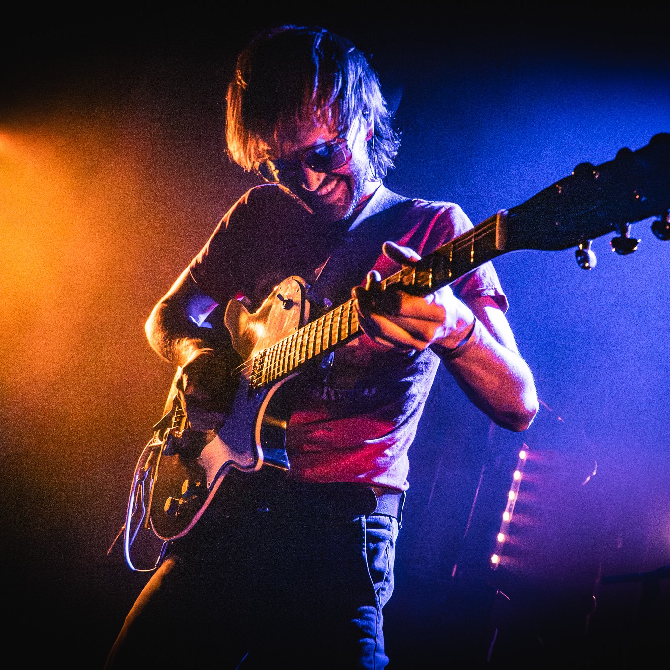 A guitarist performing on stage, wearing sunglasses. He is smiling while being focused on playing his instrument, illuminated by vibrant orange and blue stage lights