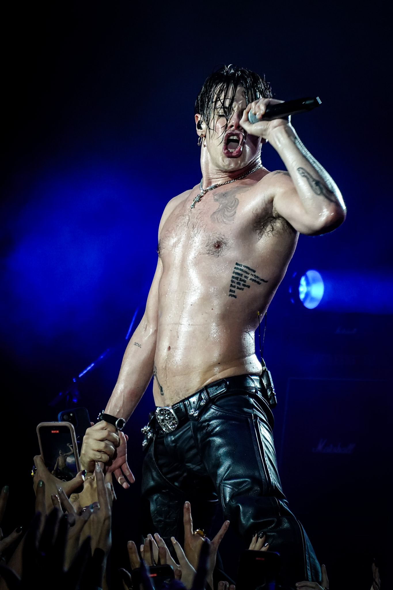 A shirtless male singer leans into the audience, performing while standing directly on top of the crowd's raised hands at a concert.