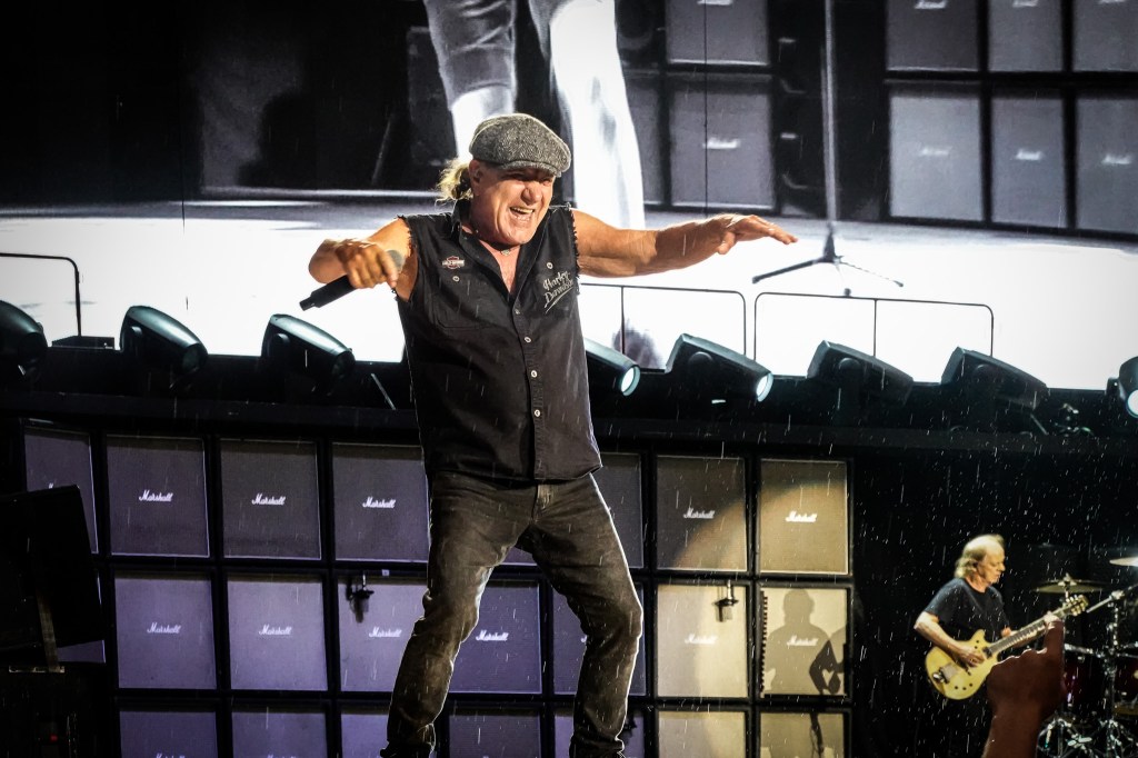 AC/DC frontman Brian Johnson performing on a rain-slicked stage during the 2026 Power Up Tour in Paris. He is wearing his iconic flat cap and a sleeveless black shirt, positioned in front of a massive wall of Marshall amplifiers, with rhythm guitarist Stevie Young visible in the background.