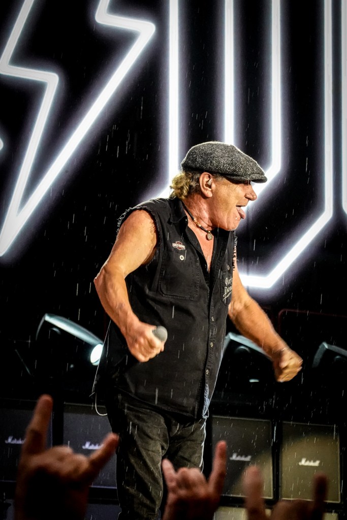 AC/DC lead singer Brian Johnson performing live in the rain at the Stade de France during the 2026 Power Up Tour in Paris. He is wearing his signature flat cap and a sleeveless Harley Davidson shirt, smiling as rain falls against a backdrop of a glowing white lightning bolt and Marshall amplifiers.