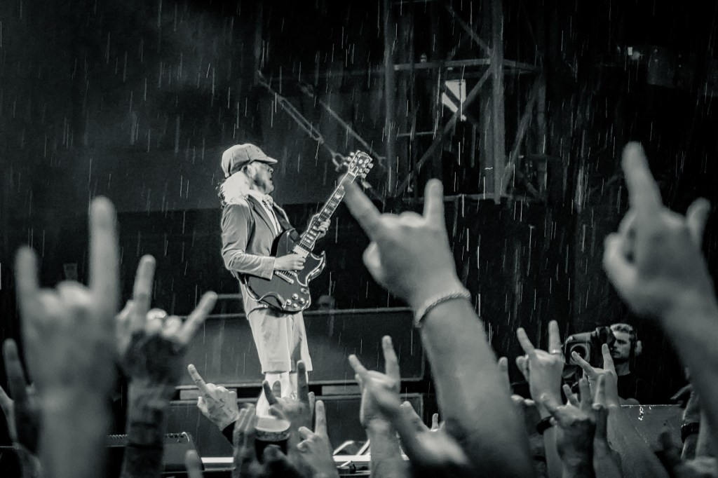 black and white photo of Angus Young performing on stage in his iconic schoolboy uniform during a heavy downpour at the Stade de France for the 2026 Power Up Tour. He is shredding on his Gibson SG while surrounded by a sea of fans raising their hands in the "rock on" hand gesture, perfectly capturing the high-voltage energy of the Paris show.