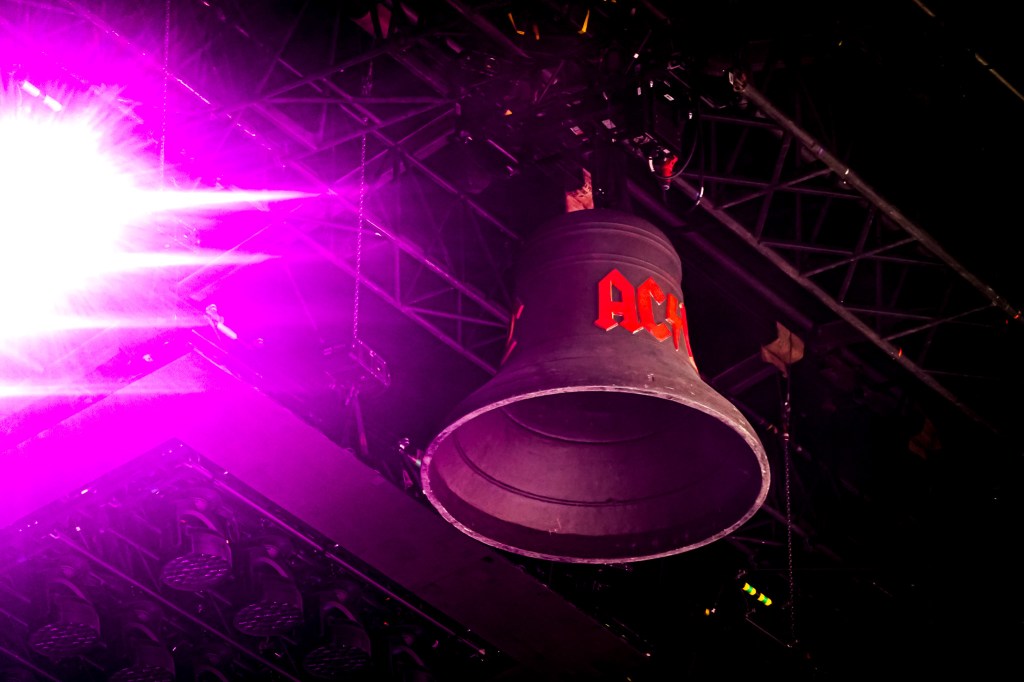 An AC/DC "Hells Bell" suspended from the stage rigging at the Stade de France during the 2026 Power Up Tour in Paris. The black bell features the red AC/DC logo and is illuminated by intense purple and pink concert lighting.