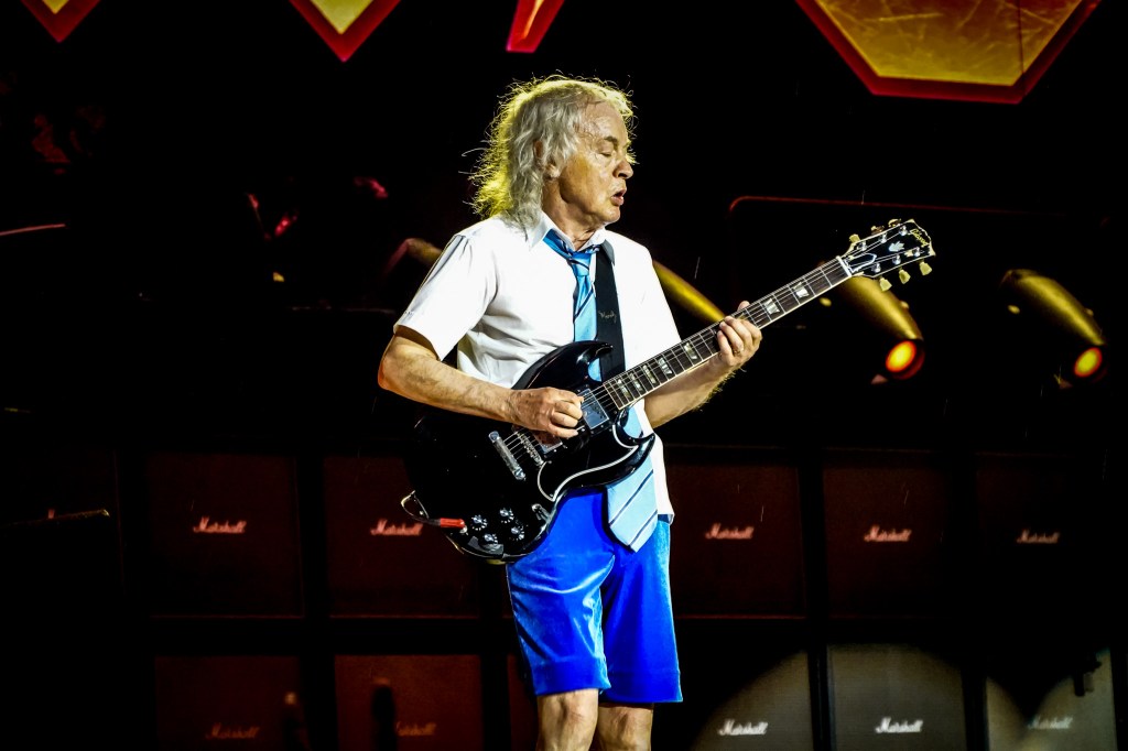 Angus Young performing a guitar solo in his signature blue velvet schoolboy uniform during the AC/DC Power Up Tour at the Stade de France in Paris. He is playing his black Gibson SG in front of a wall of Marshall amplifiers, with vibrant stage lighting accentuating the high-energy atmosphere of the 2026 live performance.