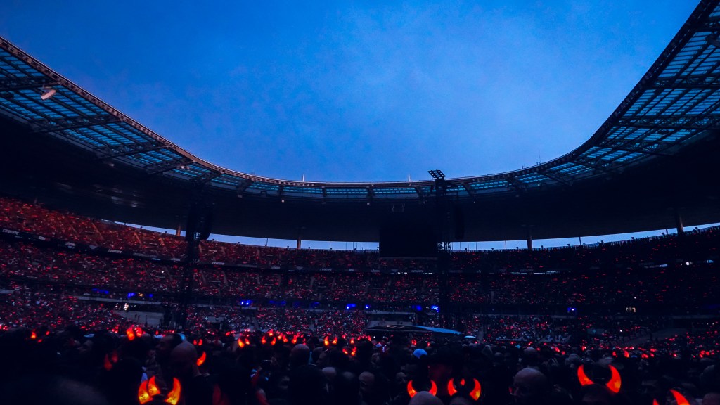 A wide-angle shot of the massive crowd at the Stade de France during the 2026 Power Up Tour in Paris. Thousands of fans fill the stadium under a deep blue twilight sky, their glowing red AC/DC devil horns creating a sea of flickering crimson lights.