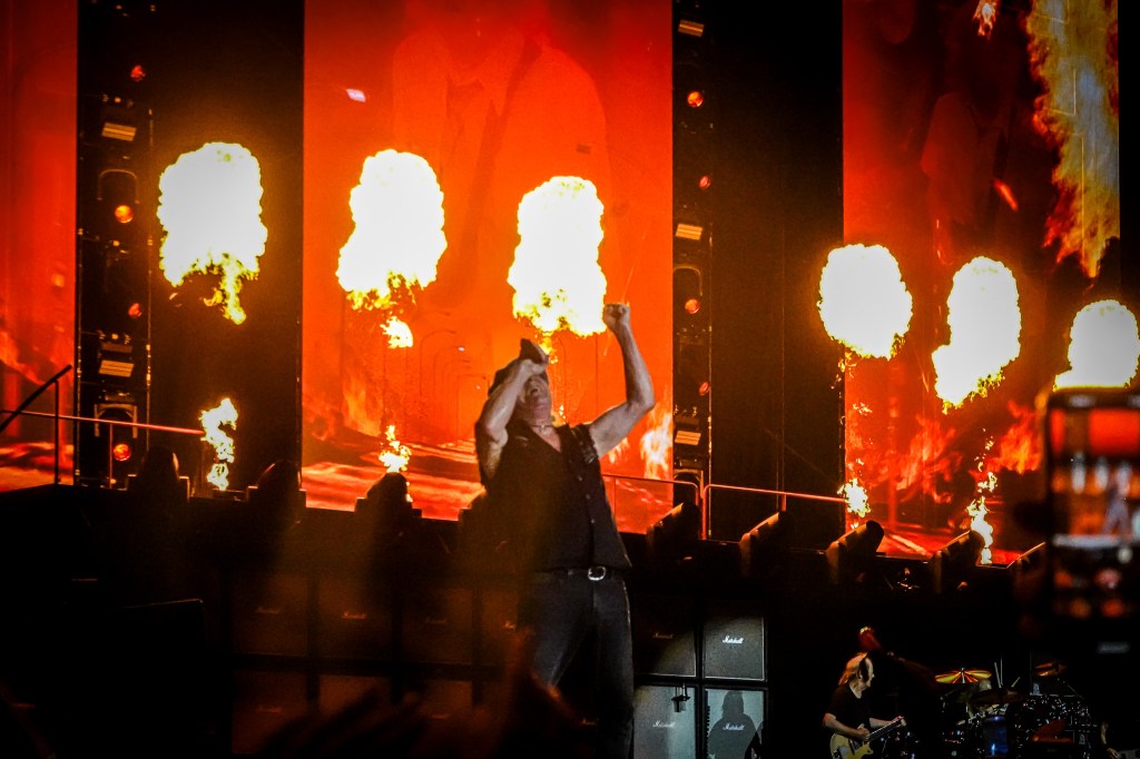 AC/DC lead singer Brian Johnson pumps his fist in front of explosive pyrotechnic fireballs during the 2026 Power Up Tour at the Stade de France. The stage is bathed in a fiery orange glow from the massive LED screens and real-time flames, creating a dramatic and high-voltage finale for the Paris audience.