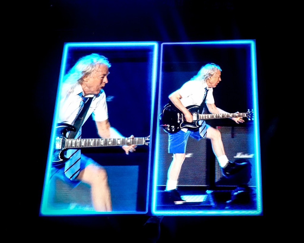 A split-screen stadium visual at the Stade de France during the 2026 Power Up Tour, featuring two dynamic shots of AC/DC guitarist Angus Young. The left frame shows a close-up of him shredding on his Gibson SG, while the right frame captures him in mid-stride across the stage in his iconic blue schoolboy uniform, both encased in glowing neon blue borders.
