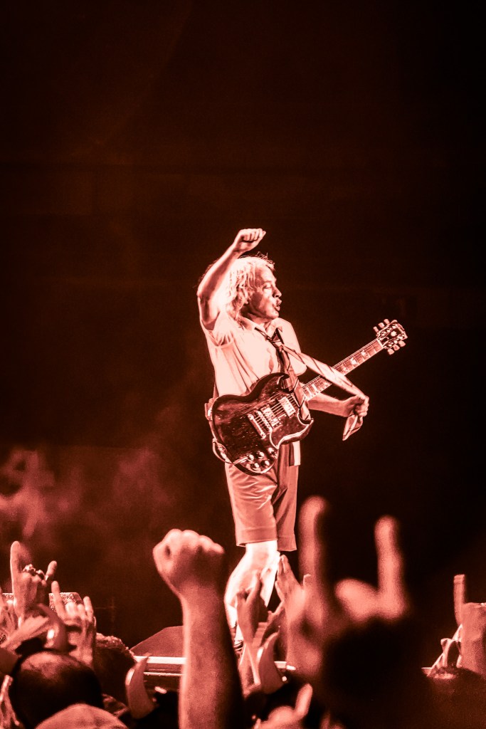 AC/DC guitarist Angus Young performing live on stage at the Stade de France during the 2026 Power Up Tour in Paris. He is wearing his iconic schoolboy uniform, pumping his fist in the air while holding his Gibson SG guitar, bathed in dramatic red stage lighting above a crowd of fans with their hands raised in the "rock on" gesture.