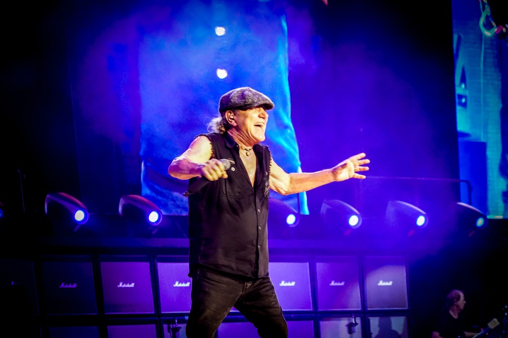 AC/DC lead singer Brian Johnson belts out a classic under intense blue stage lights at the Stade de France during the 2026 Power Up Tour. He is seen laughing and performing with arms outstretched in front of a massive Marshall amplifier stack, perfectly capturing the joy and high energy of the Parisian crowd.