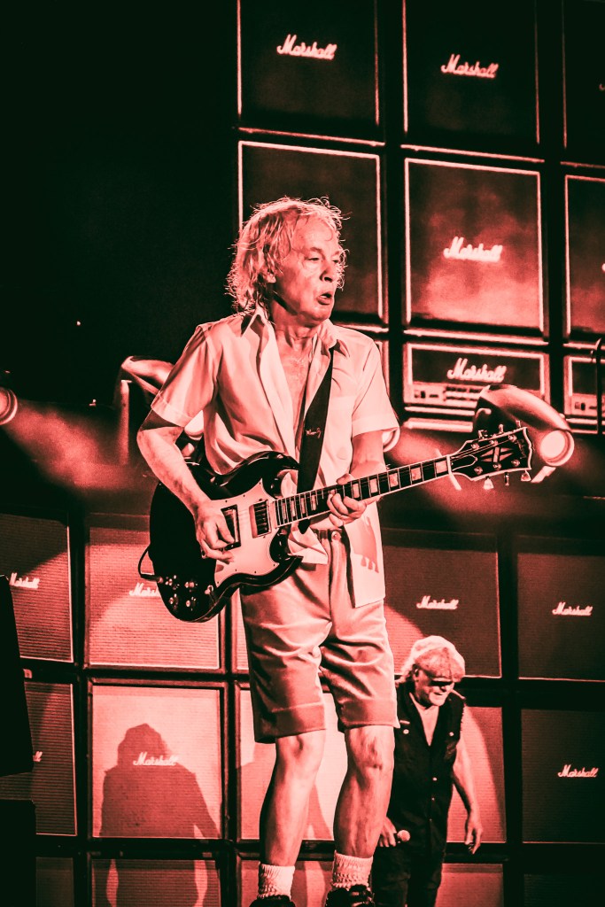AC/DC guitarist Angus Young shredding on his Gibson SG during the 2026 Power Up Tour at the Stade de France. Dressed in his iconic schoolboy uniform, he performs in front of a towering wall of Marshall amplifiers under dramatic red stage lighting, with lead singer Brian Johnson visible in the background.